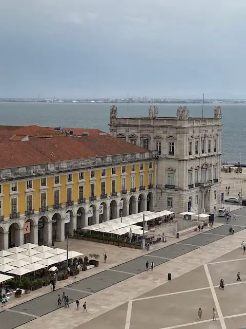 Lisbon's Majestic Praça do Coméercio, bright yellow and white arcaded building on the east side of the square.  The entrance to the  Terreiro Paço metro station is beside the L-shaped awning in the top of the photo