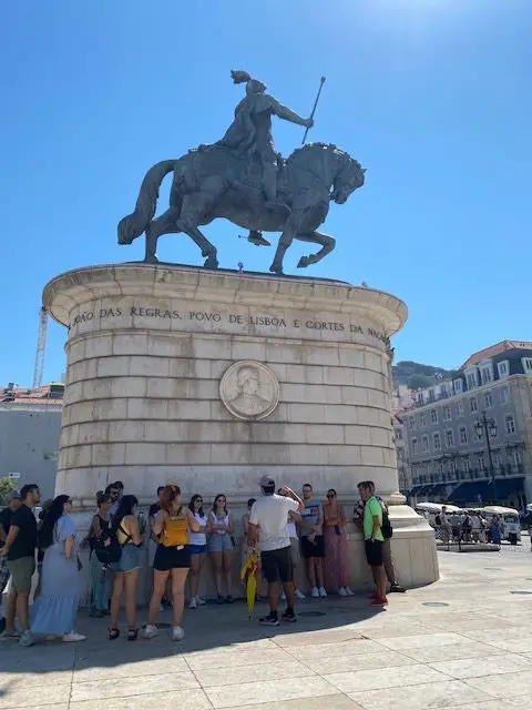 A group of tourists meets in the shade cast by the statue of Dom João I in Lisbon's Praça da Figueira