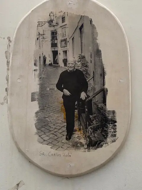 A black and white painting from Artist Camilla Watson's "Alma de Alfama" (Soulf of Alfama project, portraits of 40 elderly residents on the walls of Lisbon's Alfama nerighborhood. Here, Sr. Carlos Lola standing in front of his house.