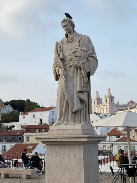 Statue of Lisbon's patron saint, São Vicente at Largo das Portas do Sol viewpoint.  Vincent holds two crows and a ship, Lisbon's symbols. An actual bird is sitting atop his head in this photo. In the background is the monastery where he lived - São Vicente de Fora monastery.