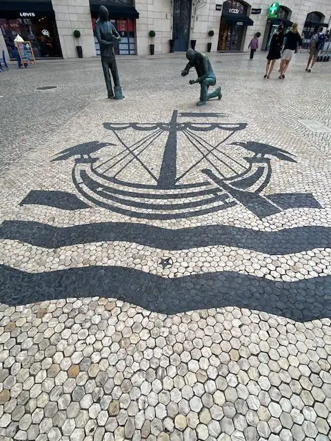 Two bronze figures, one kneeling and the other standing are a Monument to the Pavers who laid the black and white cobblestone all over Lisbon, Portugal.  THe Monumento aos Calçeteiros by Sérgio Stichini stands in front of the Avenida Palace Hotel in Lisbon.