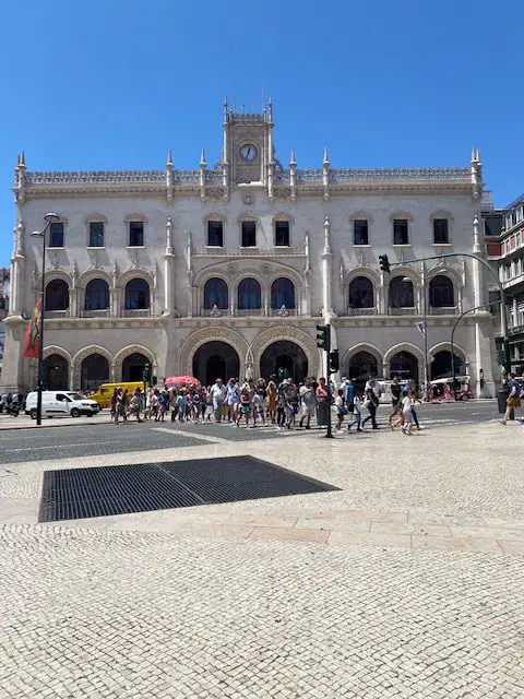 The facade of the Rossio Train Station in Lisbon , Portugal.  A large crowd is crossing the street and heading in the direction of Praça Dom Pedro IV.
